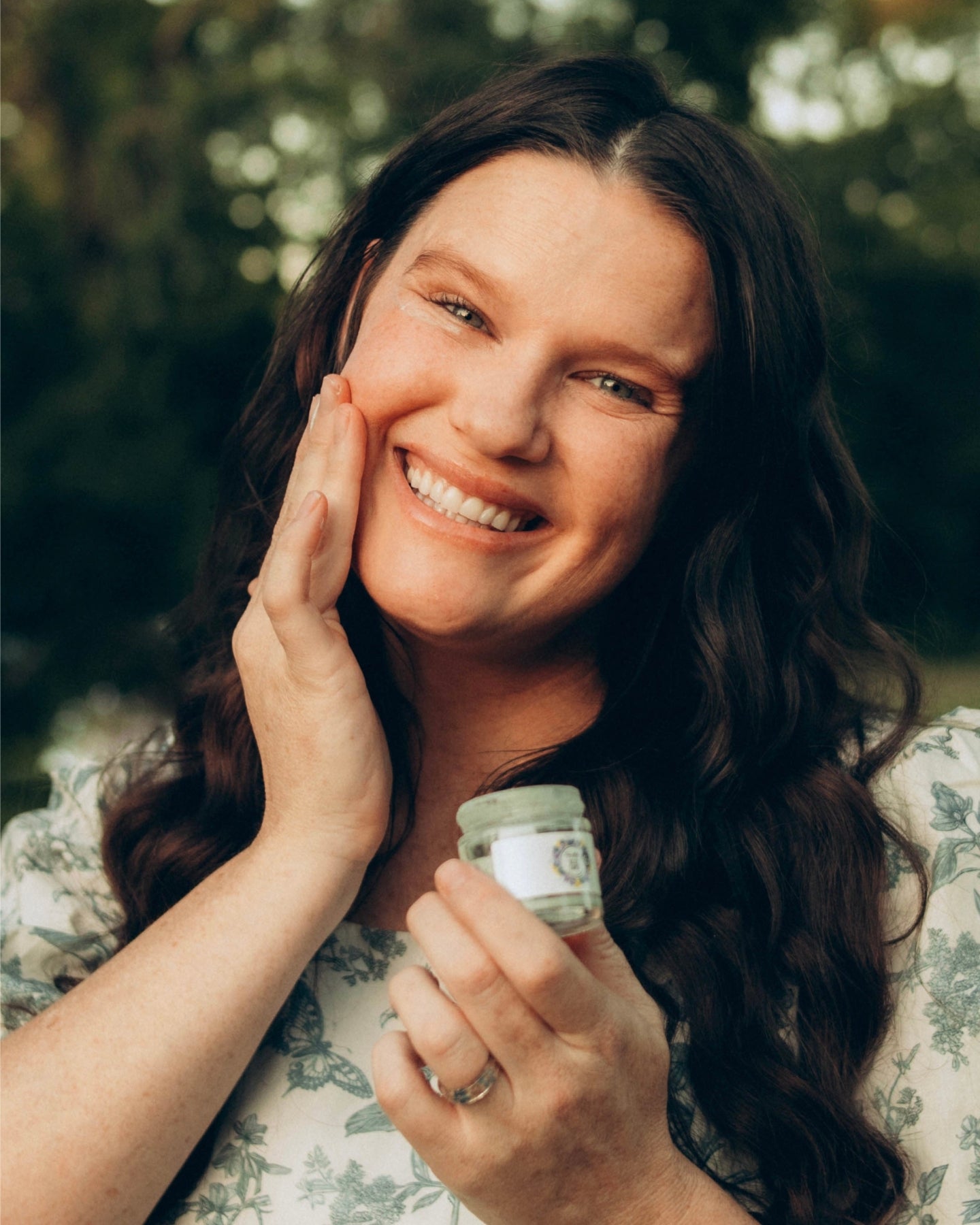 A smiling woman holds an open jar of Blue Tansy Ultra Rich Moisturizer while she massages the product onto her face.