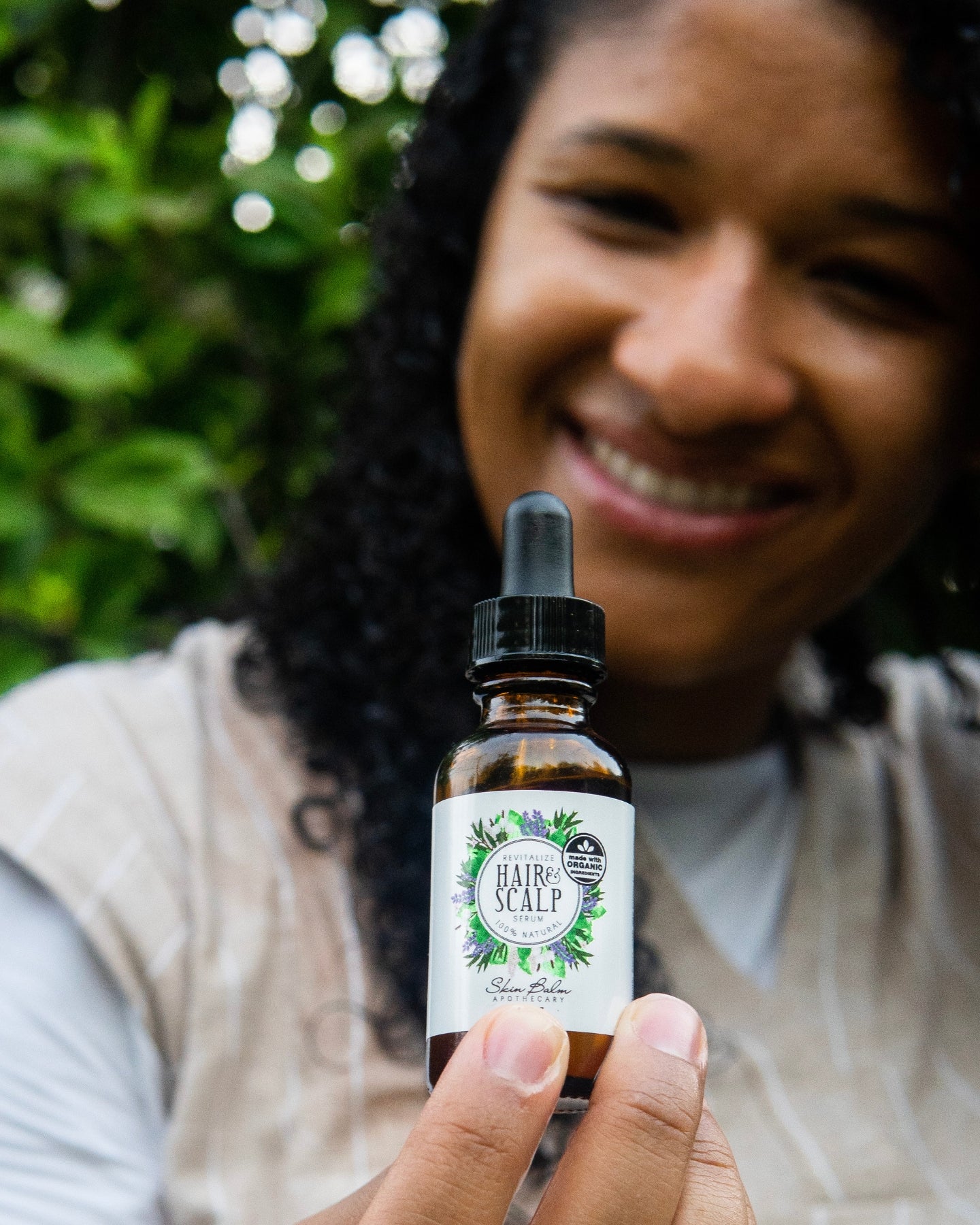 A close-up shot of the Revitalize Hair & Scalp Serum with a smiling woman blurred in the background.